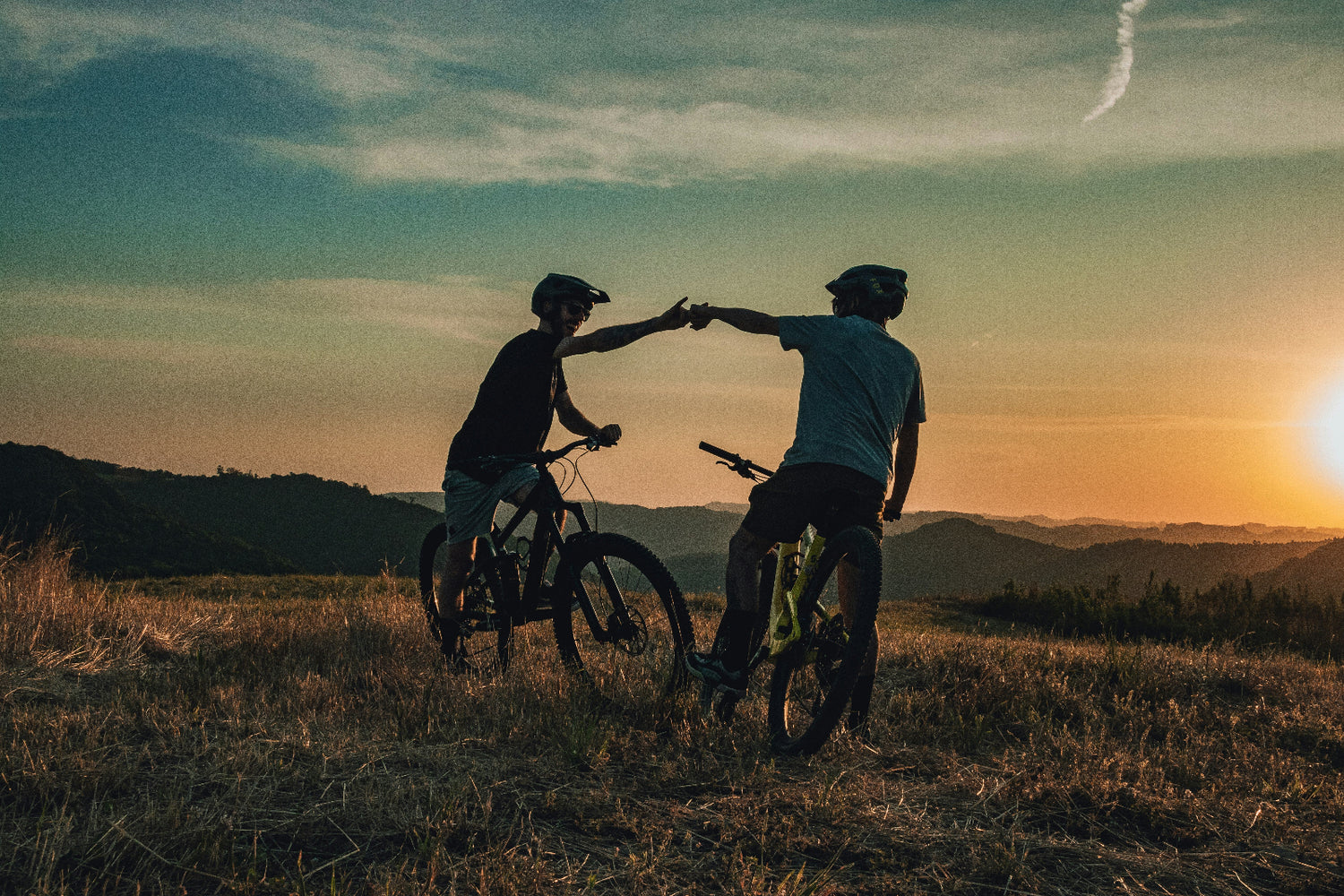 Two mountain bikers high-fiving at sunset on a scenic hilltop trail, wearing helmets and cycling gear, celebrating their ride with a panoramic mountain view.