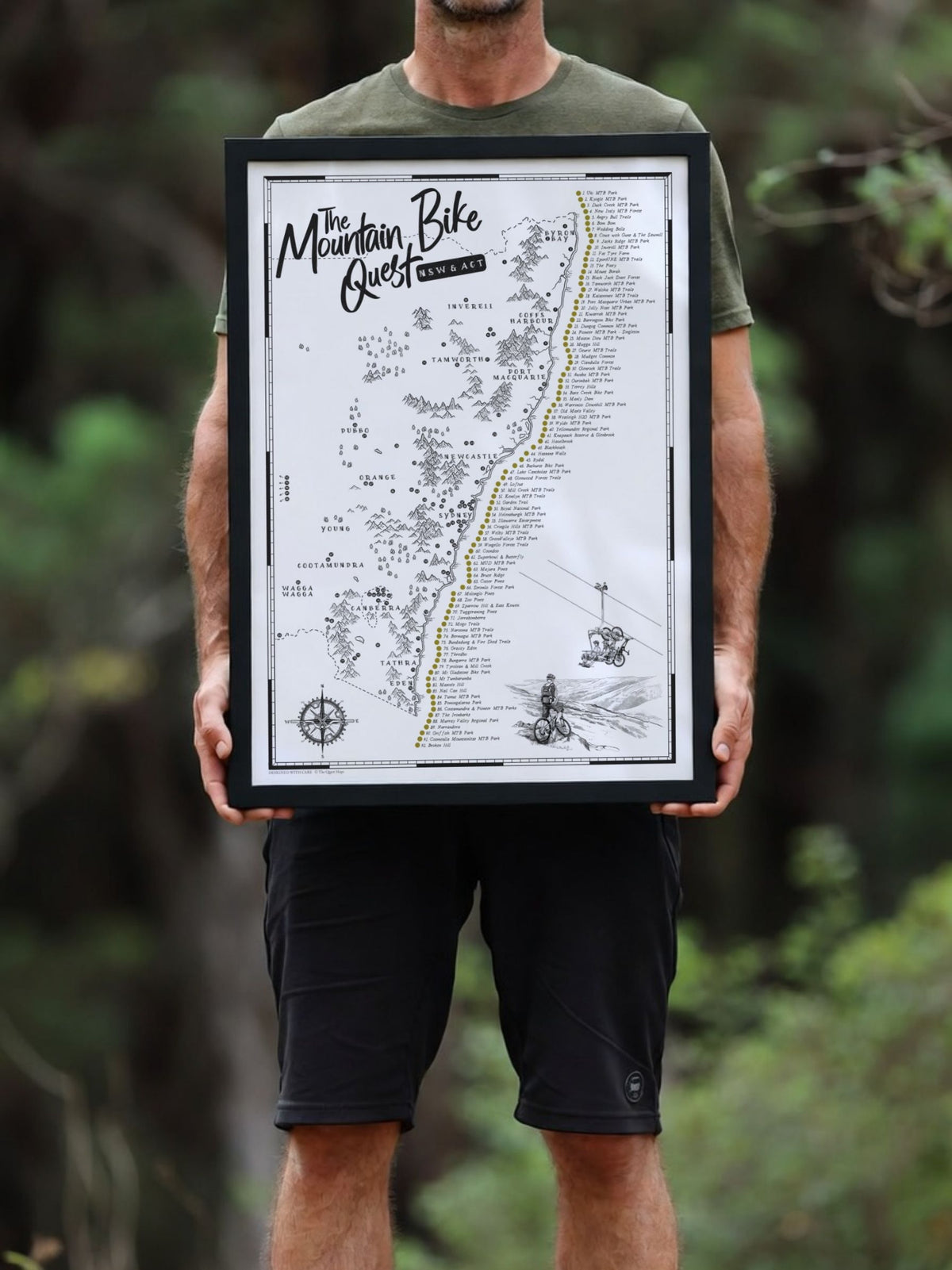 Person holding a framed NSW & ACT Mountain Bike Quest scratch map outdoors among trees
