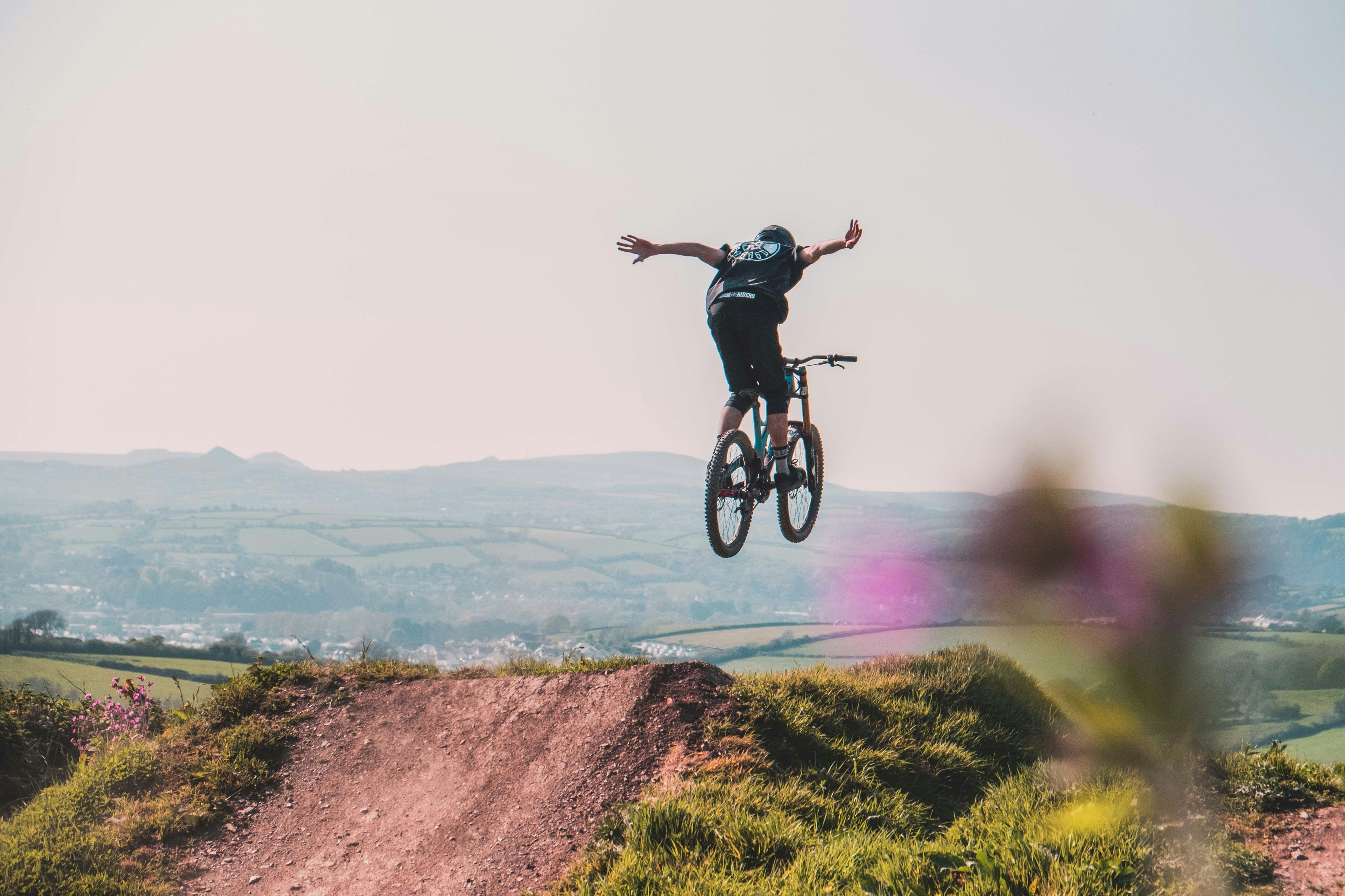 Mountain biker performing a no-hands jump on a dirt trail with rolling hills and countryside in the background, captured in mid-air.