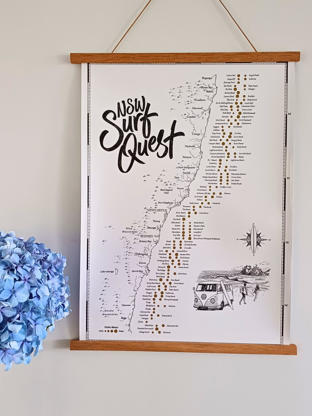NSW Surf Quest Scratch Map displayed in a wooden hanging frame on a wall, showing detailed hand-illustrated surf spots across New South Wales with gold foil scratch-off points, next to a blue hydrangea.