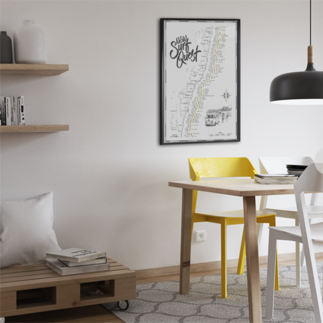 NSW Surf Quest Scratch Map framed and hanging in a modern dining room with minimalist decor, showcasing a black-and-white illustrated map of New South Wales surf spots with gold scratch-off points.